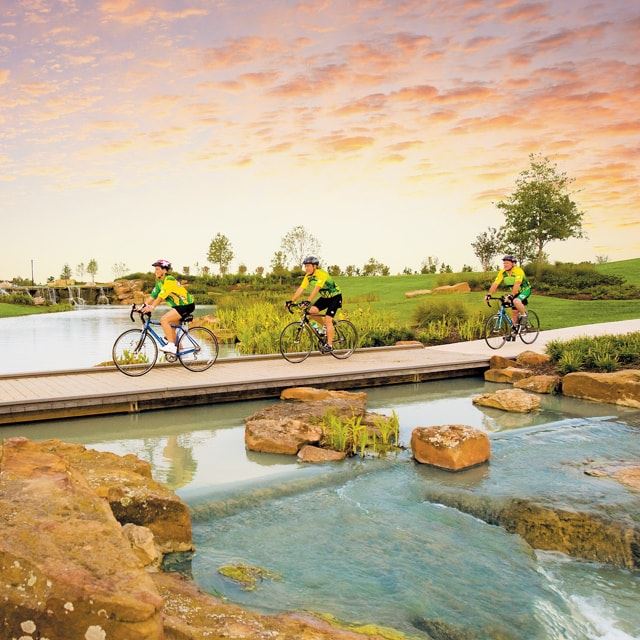 Three people riding bikes on a bridge path over a small river