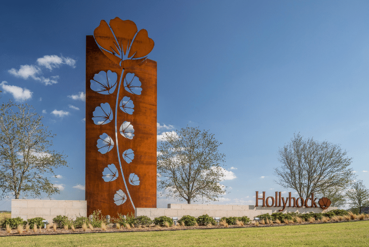 Image of the enterance to Hollyhock, a master-planned community in Dallas, Texas. You see blue skies, green grass and a small retaining wall with large letters that read, Hollyhock. To the left of the new home community name is a large steel structure showing the community's logo, the Hollyhock flower. Lining the retaining wall are green landscaped plants. Behind the structures are green trees.