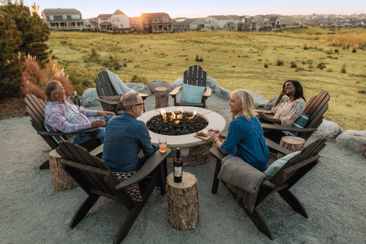 Four adults (two men and two women) sit around a fire pit in large brown Adirondack armchairs with tree stumps in between as little side tables to hold drinks. In the distance behind them you see a large field of grass with large homes in the distance past the field and blue skies above as the sun sets.