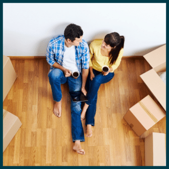 A man and woman sit next to each other, both holding a cup of coffee on their wooden floor in their new home surrounded by boxes while looking at each other smiling.